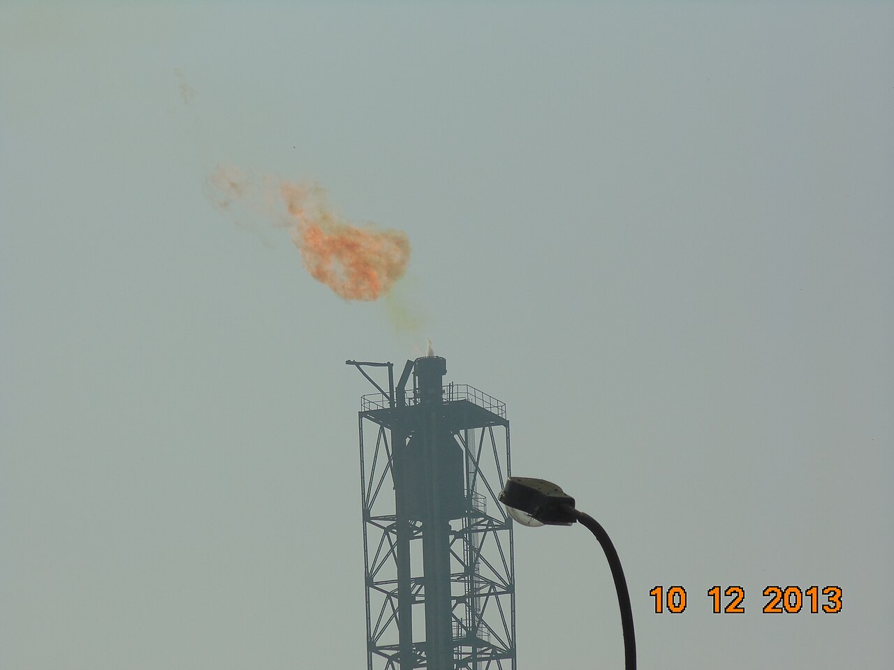 An oil refinery at dusk, with flames from a stack against the sky.