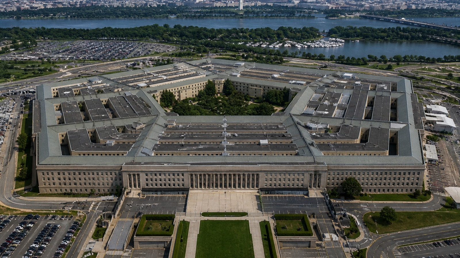 AI-generated photorealistic aerial image of the Pentagon complex in daylight, with Washington, D.C. visible in the background.
